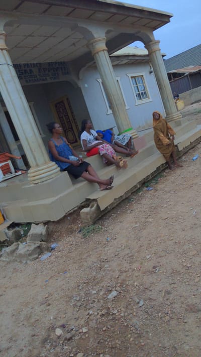Five individuals are reclining on the steps of a building. The building is identified by signage as "Chief Palace Appymiya Saa Appymiya Iswara Appymiya LGA, Nasarawa State." The building features architectural columns and a window. The surrounding environment is a dirt ground.