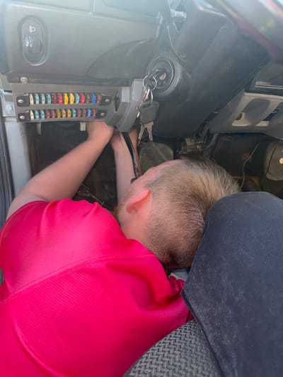 A person, appearing to be a male with short, light brown hair and a beard, is working on the fuse box of a vehicle. He is wearing a bright pink short-sleeved shirt. The fuse box is located under the dashboard, to the left of the steering column. Several fuses of various colors are visible in the fuse box. The person's head is lowered as they reach into the fuse box, holding what appears to be a tool. The steering wheel and ignition with keys are visible to the right of the fuse box. The interior of the vehicle appears to be dimly lit. There is no indication of the specific location within the city of Krasnokamsk, Russia, based on the visual information provided.