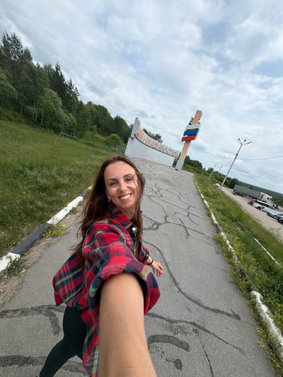 A woman in a red and black plaid shirt is taking a selfie on a cracked asphalt road. She is smiling at the camera. In the background, there is a large white sign with text in Russian that appears to say "NIKAOLAEVKA-VLADIVOSTOK". A large wooden post with three colored flags on top, red, white, and blue, is next to the sign. To the right of the road, there is a parking lot with several cars and trucks, and beyond that, a forest. The sky is cloudy and overcast.