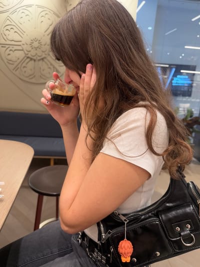 A young woman with long brown hair is seated, holding a small glass of dark liquid up to her face, possibly in a café. She wears a white t-shirt, and a black leather purse or bag with a decorative orange charm is slung over her shoulder. The table she's sitting at is light-colored wood. The location might be in Matosinhos, Portugal, though there are no strong indicators to confirm this from the image alone.