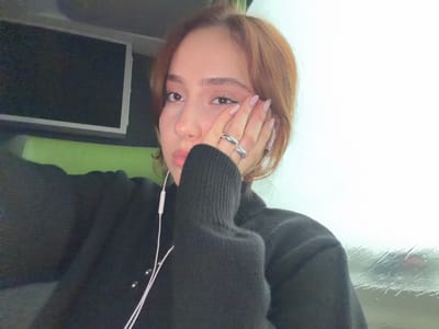 An individual is depicted in a close-up portrait inside what appears to be a vehicle in Moineville, France. The subject, a female with light brown hair, wears a dark, possibly black, long-sleeved sweater. Her left hand, adorned with two silver-colored rings on the fourth finger and light-colored manicured nails, rests against her left cheek and temple. White wired earbuds are visible, one earpiece hanging near her chest. To the subject's left, a dark rectangular screen is mounted above a bright green headrest or cushion within the vehicle's interior. To the subject's right, a window exhibits rain streaks, partially obscuring a blurred, light-toned exterior with a faint, dark-colored object, possibly a vehicle displaying red and black tones. The external conditions suggest precipitation and an overcast or foggy environment. The subject maintains direct eye contact with the camera.