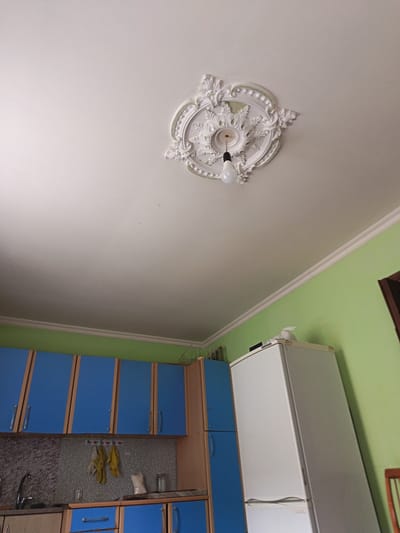 A wide-angle shot captures a kitchen area in Kasumkent, Russia. The room is brightly lit, suggesting daytime. The dominant colors are a vibrant green on the walls and electric blue on the kitchen cabinets. A white, two-door refrigerator stands against the green wall on the right. Above the cabinets, a decorative plaster medallion adorns the white ceiling, from which a single light bulb hangs. The kitchen cabinets are a mix of blue and wood-grain finishes, with silver handles. Beneath the cabinets, a granite-patterned backsplash is visible, along with a kitchen sink and faucet on the left. Some dishtowels hang on a rack above the counter. The overall impression is of a functional, lived-in kitchen space.