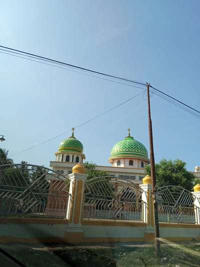 The image displays a view of a mosque with two prominent green domes topped with crescent moons, set against a clear blue sky. A yellow and white fence with intricate metalwork runs across the foreground, separating the viewer from the mosque. Several trees are visible behind the fence and surrounding the mosque. Utility wires and a wooden pole are present in the mid-ground and background. The lower portion of the image shows grass and some debris. The scene is located in the city of Meuse, Indonesia.