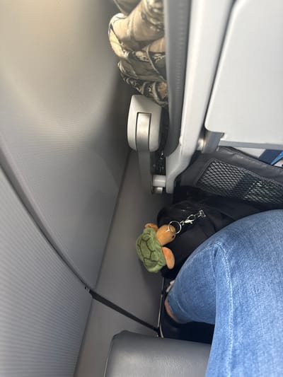 The image depicts the under-seat area of an airplane, likely during a flight or boarding process. The scene includes an airplane seat leg, a bag, and a person wearing jeans. Hanging from the bag is a plush toy turtle attached to a keychain. The flooring is a textured gray material, standard for airplane cabins. There is no information about the city of San Antonio or specific location within it.