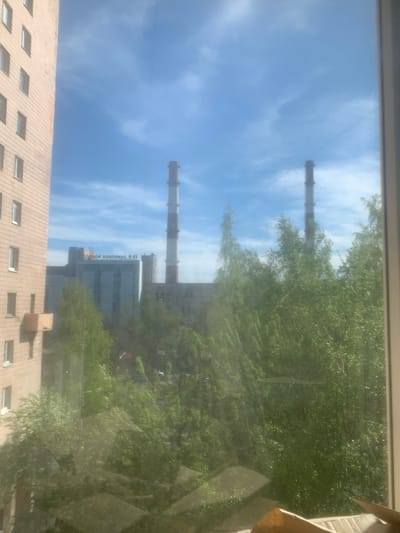A view from a window shows a mostly cloudy sky above buildings and trees. Two tall smokestacks are visible, one closer and one further away. Part of a building is visible, bearing the text "ЕЛВОВОЙ КОМПЛЕКС H49" which translates to "ELVOV COMPLEX H49." Another building with multiple windows is in the foreground. This is likely a view in or near Novoe Devyatkino, Russia.