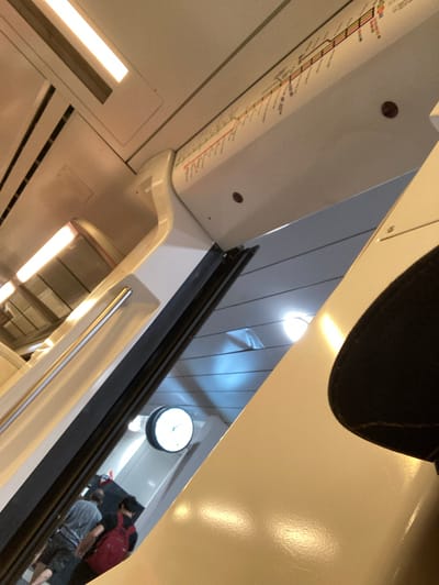 An interior view of a commuter train in Sant Cugat del Vallès, Spain. The image shows the ceiling with a fluorescent light fixture, a route map along the top of the interior wall, and the upper portion of the train's white interior. To the right, a black brim of a hat is visible in the foreground. In the middle ground, a round white clock with black hands is mounted on the wall. Below the clock, two individuals are partially visible. One person, wearing a gray t-shirt and dark shorts, is facing away from the camera. Next to them, another person wearing a red backpack and a dark top is also facing away. The environment suggests a moving train carriage with passengers.