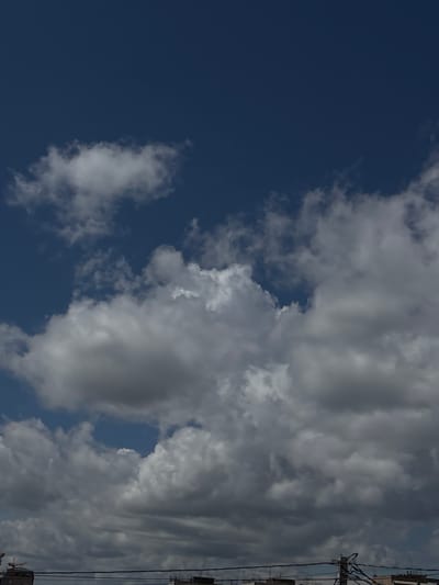 The image displays a daytime sky over Zovuni, Armenia, dominated by a dynamic interplay of clouds and blue sky. The scene is oriented vertically in the provided file, but viewed as if rotated 90 degrees counter-clockwise, it shows an expansive view from near the ground upwards.

A significant portion of the upper left quadrant of the sky is a deep, clear blue, with a solitary, wispy white cloud floating within it. Towards the center and right, the sky transitions into a dense formation of cumulus clouds. These clouds exhibit a variety of shades, ranging from bright white where they are most illuminated, to dark greys and charcoals, indicating their depth and the varying light conditions. Patches of blue sky are still visible peeking through gaps in the cloud cover.

Along what would be the horizon line at the bottom right of the image (the right edge in the current orientation), dark, silhouetted structures are visible. These appear to be parts of buildings or other man-made constructions, topped with utility poles and a network of overhead wires, suggesting an urban or semi-urban environment.

The overall lighting suggests it is daytime, likely mid-morning or afternoon, with the sun obscured or diffused by the clouds. There is no specific activity or event taking place, and no text is visible within the frame. The image primarily captures the atmospheric conditions and a glimpse of the built environment.