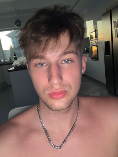 The image is a selfie of a young man with fair skin, short, tousled brown hair, and blue eyes. He is looking directly at the camera with a neutral expression. He is shirtless and wearing a silver chain necklace. His skin appears slightly flushed.

The background suggests an indoor environment, possibly a kitchen, with appliances visible. There are reflections on a shiny surface in the background, possibly windows or cabinets, showing an exterior scene with a building and sky, which appears to be dusk or dawn. The lighting is dim, with the primary light source coming from the front, casting soft shadows on his face.

There is no indication of the specific location within the city of Vanino, Russia, in the image itself.