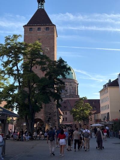 The image captures a bustling scene in Nuremberg, Germany, likely near the historic Handwerkerhof or a similar marketplace area adjacent to the medieval city walls. A prominent red-brick tower with a dark tiled, pyramidal roof dominates the left side of the frame, topped with a small bell. The tower features small, dark windows and two coats of arms. Behind the tower, a large, green dome of a building is visible.
In the foreground, a paved pedestrian area is filled with people walking and gathering. They are dressed in casual summer clothing. Several individuals are walking towards the viewer, while others are seen from behind or the side. Some people are sitting on benches or near a fountain structure at the base of the tower. A large tree with dense green foliage stands in front of the tower, partially obscuring it.
The background shows several multi-story buildings with light-colored facades and windows, typical of European city architecture. White umbrellas suggest outdoor seating or market stalls. The sky is a clear blue with wispy white clouds and contrails from airplanes, indicating fair weather. The overall impression is of a lively public space on a sunny day, with historical architecture serving as a backdrop to daily life and tourism.