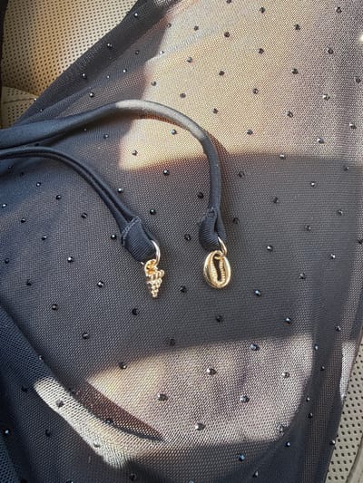 The image shows a close-up of a black sheer fabric adorned with small, dark embellishments that appear to be rhinestones or sequins. Two black drawstrings are visible, each ending in a decorative charm. One charm is gold-toned and shaped like a cluster of grapes, while the other is also gold-toned and resembles a cowrie shell. The fabric is draped over a textured surface that suggests upholstery, possibly from a car seat, and is illuminated by dappled sunlight. The scene is indoors, or in a shaded outdoor area, given the presence of the upholstery and the controlled lighting. The location is Ofrynio Beach, Greece, though no specific landmarks or contextual elements within the image pinpoint this exact location, only the general description of a beach setting can be inferred from the name. There are no people or animals present in the image.