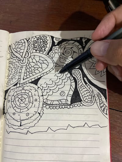 A close-up, overhead shot shows a hand holding a black pen, drawing on a lined notebook. The drawing is an abstract, doodle-like design filled with intricate patterns, swirls, and organic shapes in black ink. The notebook is open, revealing the ruled paper, and rests on a wooden surface. The pen is positioned over the drawing, as if in the process of adding more details. The image is sharp and well-lit, with the focus on the drawing and the hand. There are no discernible landmarks or features that would indicate a specific location in Los Velásquez, Venezuela.