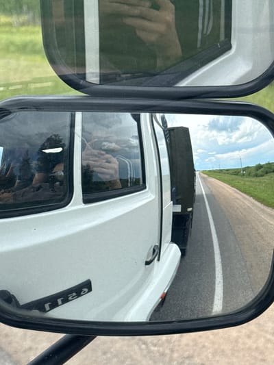 This media file shows a reflection in a truck's side mirror. The mirror captures a view of a road stretching into the distance, with a grassy field and blue sky with clouds on either side. A truck is visible in front of the viewpoint, also reflected in the mirror. The scene appears to be set during the daytime, with bright sunlight indicating clear weather.
The truck's exterior is white. Text that appears to be "JJaz" or a similar reversed word is visible on a black strip on the truck's body. Inside the truck, the reflection in the window shows a person's hand holding a phone, suggesting the driver is using or about to use their phone. Another reflection in a lower window appears to show a person wearing a dark top and glasses, possibly the driver.
The location context provided is Қызылжар ауылдық округі, Kazakhstan. This suggests the image was taken on a road in this rural area of Kazakhstan. The overall impression is a snapshot of a moment during a drive, possibly capturing a mundane activity like checking a phone, juxtaposed with the open road and natural scenery.