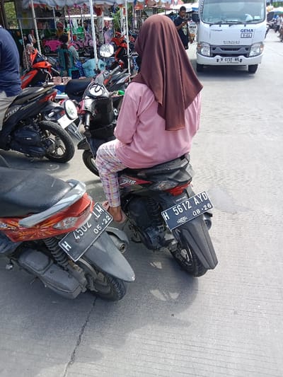 The image shows a woman wearing a pink shirt and a brown hijab, sitting on a black motorcycle with a red frame. The motorcycle is parked on a concrete surface. Another motorcycle is visible in the foreground, with a red frame and a black seat. In the background, a white van with "GEMA T-01" written on the back is parked further away. The scene is located in Kendal, Indonesia, at the coordinates -6.932 latitude and 110.203 longitude. The woman is sitting upright on the motorcycle with her legs extended, her right foot on the ground and her left foot on the motorcycle's footrest. There are other people walking around in the background.