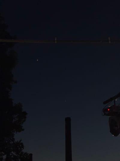 The image shows a dark sky with two distinct points of light, likely stars or planets. In the foreground, on the right side of the image, a dark object with a red light is visible, possibly a piece of lighting equipment. A dark, slender vertical structure, resembling a chimney or tower, is positioned in the lower center of the frame. On the left side, a dark silhouette of foliage from a tree is visible. A horizontal line, possibly a power line or cable, stretches across the upper portion of the image. The scene appears to be an outdoor, urban or semi-urban environment at dusk or night.