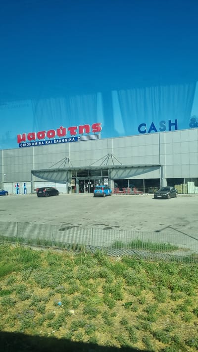 The image shows the "Masoutis" supermarket and "Cash & Carry" store located in Larissa, Greece, as indicated by the signage. The building is a large, modern structure with a grey facade and large glass windows. Above the entrance, the red "Masoutis" logo is prominently displayed with the Greek text "ΟΙΚΟΝΟΜΙΚΑ ΚΑΙ ΕΛΛΗΝΙΚΑ" below it, meaning "Economical and Greek". To the right, the blue text "CASH &" is visible. The parking lot in front of the store is paved and features several parked cars, including a black sedan on the right and a black SUV on the left, with a smaller blue car parked further back. Shopping carts are visible near the entrance. The surrounding environment includes a metal fence and a grassy area with scattered plants. The sky is a clear, bright blue, suggesting a sunny day.