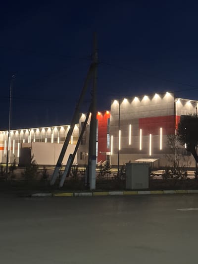 The image captures the facade of a modern building at night, illuminated by numerous lights. The building features a prominent red and white color scheme, with a series of horizontal light fixtures arranged across its upper section. Above these, a line of upward-pointing spotlights creates a dramatic effect. Trees are visible along the side of the building, casting shadows. To the right of the building, a paved road with painted lines is evident, bordered by a curb with yellow and white markings. A utility box is also visible near the building. In the foreground, a power line structure with diagonal supports and wires cuts across the frame, silhouetted against the dark blue night sky. The scene suggests an urban or commercial setting in Petropavl, Kazakhstan, after dark. There is no visible text in the image.