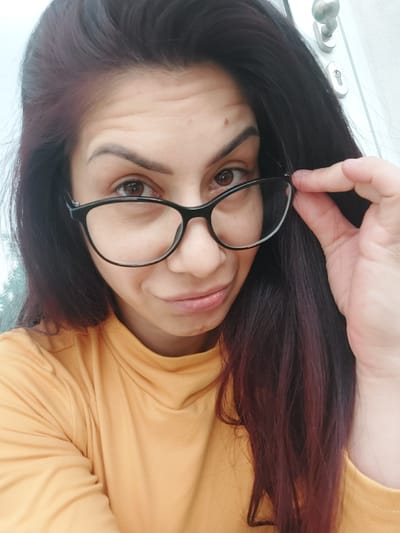 A woman with dark hair and glasses poses for a selfie. She is wearing a yellow turtleneck shirt. Her right hand is touching her glasses. In the background, a white door with silver hardware is visible.