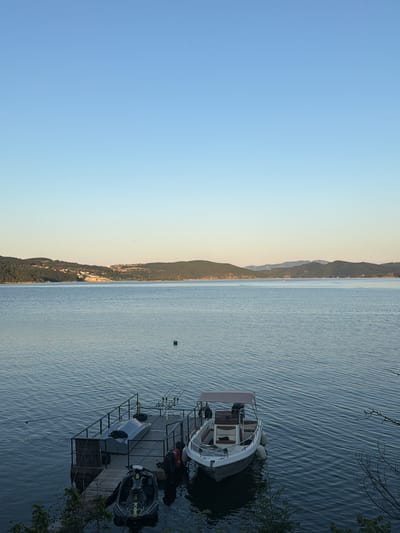 A serene lake view is captured with rolling green hills in the background under a clear, pale blue sky. The water is calm with gentle ripples, reflecting the muted colors of the landscape. In the foreground, a small wooden dock extends into the water. Moored to the dock are two watercraft: a jet ski and a small motorboat with a canopy. The motorboat is white with brown accents and has two fenders attached to its side. Fishing rods are set up on the dock. The scene appears to be in the city of Zvanika, Bulgaria.