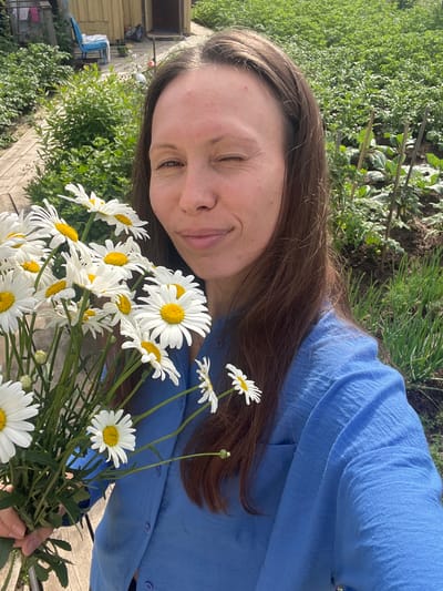 This is a portrait of a woman holding a bouquet of daisies. She is looking towards the camera with a slight smile, her eyes partially closed. Her long, brown hair is down, framing her face. She is wearing a blue collared shirt. The background is outdoors, with greenery and some gardening beds visible, suggesting a garden or yard setting. There are also some wooden structures and a blue chair in the background, potentially indicating a rural or suburban location. The lighting suggests it might be daytime, with the sun casting shadows. The overall mood appears relaxed and cheerful. The image appears to have been taken in Pigozino, Russia. There is no visible text in the image.