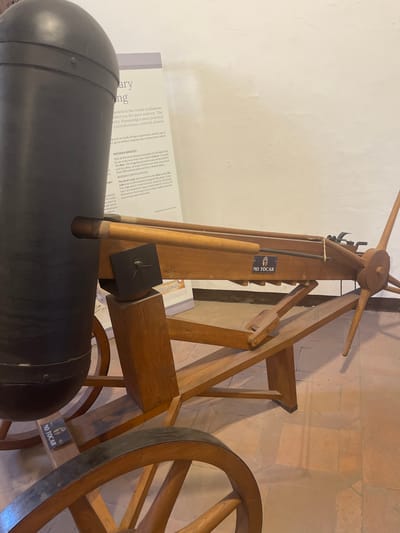 An exhibit display shows a large black cylindrical object on a wooden wheeled cart. The cart has a wooden framework with two large wheels. A wooden mechanism with an arm and a propeller-like element extends from the back of the cart. A sign with "NO TOCAR" (DO NOT TOUCH) is visible on the wooden structure. Behind the cart, a vertical informational plaque describes ancient Roman engineering achievements. The floor is tiled with terracotta-colored squares. The wall behind the exhibit is plain and cream-colored. This exhibit is located in Toledo, Spain, within the Alcázar of Toledo, which houses the Army Museum (Museo del Ejército).