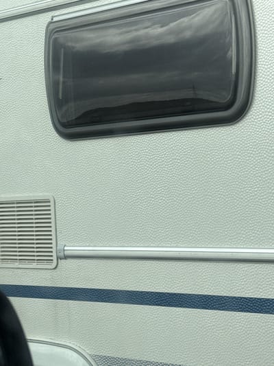 The image shows a close-up view of a white, textured caravan exterior. A dark-tinted, rectangular window with a black frame is visible in the upper portion of the image, reflecting a cloudy sky. To the left of the window, lower down, is a rectangular ventilation grill. A horizontal silver trim piece runs across the caravan, just below the window and above the ventilation grill. Below this trim, a dark blue stripe runs horizontally across the white surface. The lower part of the image shows the bottom edge of the caravan, including a silver trim and a curved silver frame, possibly for another window or door. A black object, possibly a steering wheel or part of a seat, is partially visible in the bottom left corner, suggesting the photograph was taken from inside a vehicle. The scene is not definitively located in Meppel, Netherlands, as there are no identifying landmarks or context provided within the image.