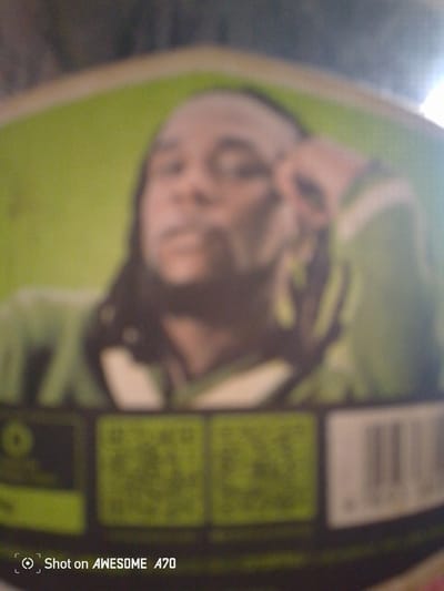 The image is a close-up, low-resolution photograph of a person. The person appears to be a male with dark skin and dark hair, wearing a green garment with white stripes. The background is blurred and appears to be a green surface. At the bottom of the image, there are two QR codes and a barcode, as well as text that reads "Shot on AWESOME A70". The image lacks sufficient detail to determine the exact location or any specific actions or interactions. The setting is not identifiable as Magado, Nigeria.
