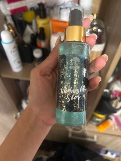 A person is holding a bottle of "fiorevita Glitter Body Mist Midnight Star" in front of a shelf filled with various personal care products. The bottle is partially filled with a shimmering turquoise liquid. The person's hand, with long acrylic nails, is visible holding the bottle. The background shows a cluttered shelf with items like a white spray bottle, a yellow bottle, a black bottle with an orange lid, and other unidentifiable products. The overall environment appears to be a bathroom or vanity area. The image does not provide any information about the city of Perushtitsa, Bulgaria.