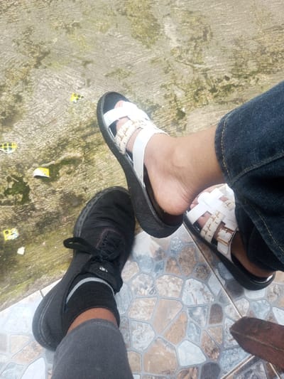 This media file presents a top-down view of two individuals' lower legs and feet in Batulawang, Indonesia, under bright daytime lighting. The scene features two distinct ground surfaces.
On the left, one person wears a black lace-up shoe with a black sock and dark grey or black pants. Their foot is positioned on a coarse, light grey concrete surface, which exhibits small patches of green growth, possibly moss or algae, and scattered tiny yellow and white debris.
On the right, another person wears white open-toed sandals with a prominent black sole. These sandals feature white straps with an intricate woven pattern in a beige or light brown color, secured with a gold-colored buckle. This individual is wearing dark blue jeans. Their foot rests on a patterned tiled floor, composed of irregularly shaped pieces in various shades of grey, brown, light blue, and white.
The presence of two different ground textures suggests a transitional area, such as a threshold between an outdoor concrete path and a tiled indoor or patio space. A small, reddish-brown object is partially visible on the tiled floor in the bottom right corner. No specific activity is observable beyond the subjects' presence, and no text is visible within the image.