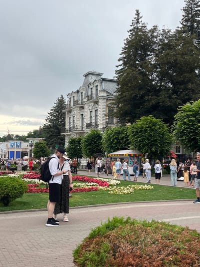 The image depicts a street scene in Kislovodsk, Russia, likely taken during the daytime under overcast skies. A large, ornate white building with multiple balconies and a distinctive mansard roof dominates the background. In the foreground, a couple stands close together on a brick-paved walkway, facing away from the camera. The man wears a white shirt, black shorts, a black backpack, and black sneakers. The woman wears a patterned skirt and sandals. They appear to be looking at a phone.
To their left, a well-maintained flower bed displays a vibrant mix of pink and white flowers, bordered by green grass. Beyond the flower bed, a crowd of people are visible, walking and gathered near a small, colorful food stall or kiosk. Trees with lush green foliage line the street, providing shade. The overall atmosphere appears to be that of a public gathering or a bustling urban square.