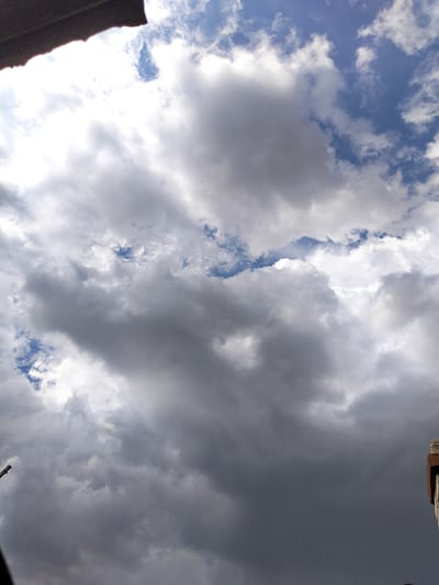The image shows a view of the sky, dominated by a dramatic display of clouds. The clouds are varied in texture and color, ranging from bright white and luminous where the sun is shining through, to dark, ominous grey in other areas. Patches of blue sky are visible between the clouds, suggesting a dynamic weather pattern. The upper left corner of the image is partially obscured by what appears to be the edge of a dark, textured surface, possibly a roof or awning. In the lower right corner, a section of a building's exterior is visible, made of brick and a darker material, possibly a chimney or part of a wall. The bottom left shows the tip of a dark pole, possibly a streetlamp or antenna. The scene is likely an outdoor urban environment in Luhansk, Ukraine, given the context provided. There are no people or animals visible in the image. The lighting suggests it is daytime, with the sun intermittently breaking through the cloud cover.