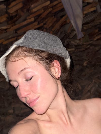 The image shows a close-up of a woman's face and shoulders. She is wearing a gray cap with a white trim and has her eyes closed, with her head tilted down. Her hair is wet and appears to be pulled back. She has light skin and appears to be in a sauna or steam room, indicated by the steamy appearance of her skin and the rustic wooden background. There are no visible landmarks or indications of the city of Maksimovo, Russia, in the image.