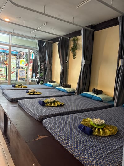 The image displays the interior of a massage establishment located in Ban Ko Li Pe, Thailand. Six individual massage stations are arranged consecutively on a raised, dark brown wooden platform. Each station is equipped with a dark blue mattress featuring a golden geometric pattern, a light blue pillow, and a small, dark brown cylindrical bolster pillow. Five of the visible stations are accessorized with a decorative arrangement at the foot of the bed, comprising a rolled dark blue towel, a folded green and gold patterned fabric, and a white artificial lotus flower. Grey fabric curtains hang from a white ceiling-mounted track system, providing privacy screens along a light yellow-painted wall. Fluorescent light fixtures are visible on the white ceiling. A large glass storefront on the left side of the room offers a view of an exterior street or pathway, where various commercial signs, posters, and merchandise from adjacent businesses are discernible. An artificial green vine plant is suspended on the yellow wall between two curtain sections. No individuals are present in the scene.