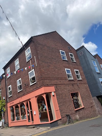 The image shows a brick building in Knutsford, United Kingdom. The building is multi-story, with a dark gray addition on top. It has numerous white-framed windows. The front of the building features a pink storefront with arched windows, labeled "BATH HOUSE." A string of red, white, and blue pennant banners is draped across the facade. A narrow sidewalk with a yellow line runs along the side of the building. The sky is partly cloudy.