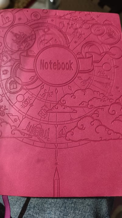 The image shows the cover of a pink notebook. The cover is embossed with a circular design featuring various icons and patterns. In the center of the circle, the word "Notebook" is embossed in capital letters within a smaller circular frame. The notebook is placed on a surface that appears to be blue denim fabric. A pink ribbon bookmark is visible hanging from the top edge of the notebook. The notebook cover is textured and the embossed elements cast subtle shadows, indicating a light source from above and to the right. There is no indication of the location being in Malaya Shil'na, Russia, or any specific city.