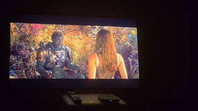The media file shows a scene from a movie or TV show displayed on a screen. In the foreground, on a dark surface, are two objects: a black remote control and a small, rectangular black device with a white light.
The screen itself displays a vibrant and colorful scene. On the left, a Black man is shown wearing an ornate, futuristic-looking green and gold armor with shoulder pads. He has dark, braided hair and is looking towards the right with an expressive facial expression. His left hand is extended, and his right hand is holding something near his waist.
On the right side of the screen, a woman is seen from behind. She has long, brown hair and is wearing a white top. She is facing the man and appears to be engaged in a conversation or interaction with him.
The background behind the two individuals is a lush, alien-like landscape. There are tall, dry grasses, purple and pink flowering plants, and what appear to be white, round seed pods or fruits. Several small, blue butterflies are also visible, adding to the surreal and fantastical atmosphere. The lighting suggests it might be daytime, possibly with a golden hour glow due to the warm tones in the foliage. The overall mood of the scene appears to be one of discovery or emotional exchange.
There is no visible text within the media file. The setting appears to be an outdoor, natural environment, but with elements that suggest it is not Earth. Given the provided location context of Udachny, Russia, this scene is unlikely to be from that specific geographical location but rather a fictional setting.