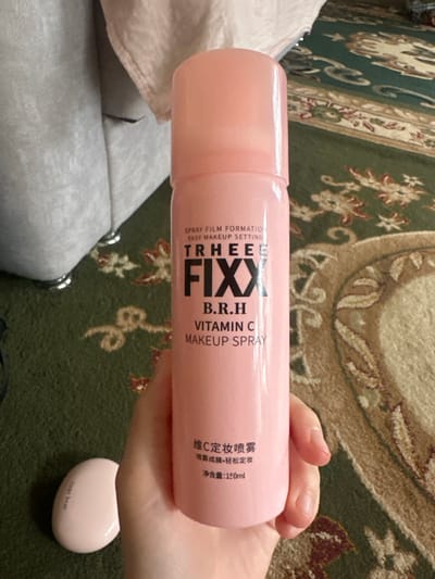 A person's hand is holding a pink aerosol can of makeup setting spray. The can has black text that reads "TRHEER FIXX B.R.H VITAMIN C MAKEUP SPRAY" and "维C定妆喷雾 喷雾成膜·轻松定妆 净含量:150ml". A small pink object with the text "Gege bear" is visible in the lower right corner. The background is a carpet with a floral pattern in green and tan. The lighting appears to be indoor, with no specific time of day or weather cues. The overall impression is a close-up product shot.