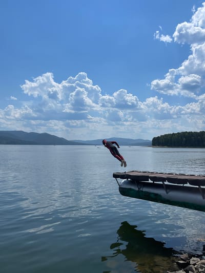 In Divnogorsk, Russia, a person wearing a red and black wetsuit is captured mid-air as they jump from a wooden dock over a calm body of water. The dock, made of wood planks supported by what appears to be a green cylindrical float and tire fenders, extends into the frame from the right. The person is positioned over the water, with their body contorted in a dynamic pose, suggesting they are in the process of diving. The background features a vast expanse of water, a tree-lined distant shore with rolling hills, and a bright blue sky filled with fluffy white clouds. The water reflects the sky and surrounding landscape, creating a serene atmosphere. Several small boats are visible in the distance on the water. The overall scene depicts an active moment in a picturesque natural setting.