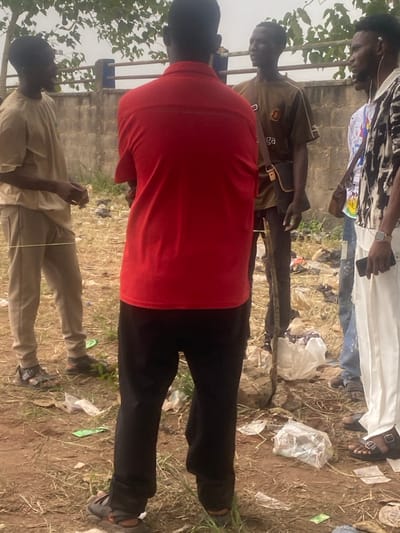 This image captures an outdoor scene in what is identified as Igbesa, Nigeria, showing a group of at least five men standing on dirt ground littered with plastic bags and other debris. The perspective is from behind a man in the foreground wearing a red short-sleeved shirt and black pants.
To his left, another man in a light brown traditional outfit (possibly a caftan) and sandals is visible, looking down with his hands seemingly engaged in a delicate activity, possibly manipulating a thin string or small object. Directly behind the red-shirted man, another individual wears a dark brown or olive green short-sleeved shirt, potentially with a logo or text like "Balenciaga" (though unclear), and dark pants, with a strap across his chest. Further back and to the right, a man in a white long-sleeved shirt or robe and dark pants with sandals is present, wearing earphones. Another person in a patterned shirt is partially visible on the far left.
The ground is dry, dusty earth with sparse vegetation and numerous pieces of white and clear plastic litter. A low concrete wall or barrier runs across the middle ground, separating the subjects from the background. Trees and foliage are visible on the left side of the frame. The lighting suggests it is daytime, with bright, diffused light indicating an overcast sky or bright ambient conditions, as no harsh shadows are apparent. The general atmosphere is calm, with the individuals appearing to be observing or interacting in a casual setting. No discernible text is clearly visible on any object or surface in the image beyond potential branding on a shirt.