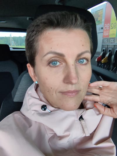 A selfie taken inside a car shows a woman with short, dark hair and blue eyes. She is wearing a light pink jacket. The background shows a gas station with a price display. The scene is likely in Фаніпальскі сельскі Савет, Belarus.