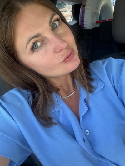 A woman with brown hair and blue eyes takes a selfie in the back seat of a car. She is wearing a blue collared shirt and a silver necklace. In the background, a deflated inflatable tube is visible. The scene is likely within the city limits of Lavans-lès-Saint-Claude, France, given the car's interior and the presence of an inflatable tube suggesting outdoor activities.