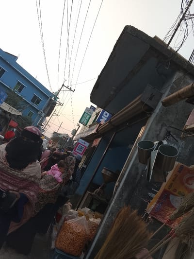 The image depicts a busy street scene in Tarabo, Bangladesh, captured with an upward and rightward tilted perspective. The pale, hazy sky indicates either early morning or late afternoon light.
On the left, a multi-story building with a light blue exterior and multiple rectangular windows is visible. Numerous overhead power lines crisscross the upper sky, connected to utility poles extending down the street.
In the mid-ground, a group of people, primarily women, are positioned along the street. The closest individual on the left wears a dark face-covering (niqab/hijab) and a patterned shawl in beige, red, and brown tones. Immediately behind her, another woman carries a small child, possibly a baby or toddler, dressed in pink, slung on her back/side. Other individuals with head coverings are visible further down the street.
To the right, the side of a concrete building forms a shopfront. An extended concrete ledge serves as an awning. Visible on the building are signs, including a blue sign with "ON" text, a green sign, and a white sign displaying a red QR code for "Nagad Pay." A partially closed roller shutter is present above the shop entrance. Inside the shop, a large, metal, funnel-shaped object is discernible. On the exterior wall, two silver-colored metal cups or buckets hang from a fixture. Lean against the wall are several brooms made of natural bristles with red-wrapped handles. At the base of the wall, clear plastic bags filled with what appear to be fried snacks or grains are displayed. A large, colorful sack with Bengali text is partially visible on the far right.