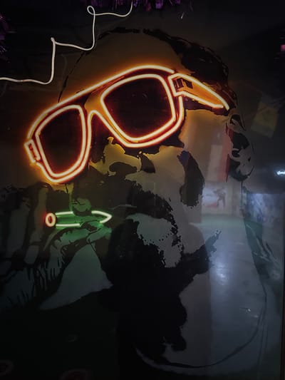 The image depicts a poster or artwork featuring a stylized portrait of a person. The face is rendered in black and white with an effect that suggests it is worn or weathered. Superimposed on the face are neon light representations of sunglasses in a bright orange-yellow color and a cigarette in green, with the tip burning in red. The image is likely taken indoors. There is no verifiable information in the image to suggest the location is Linghe, China.