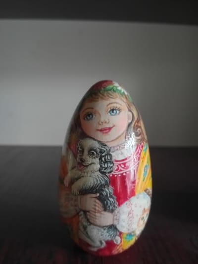 A close-up, head-on, eye-level shot shows a matryoshka doll, an egg-shaped wooden doll, hand-painted with a young girl holding a black and white dog. The girl has blue eyes, blonde hair, and rosy cheeks. She is wearing a red dress adorned with white trim and a yellow shawl. The dog is fluffy with dark patches around its eyes and on its ears. The doll is on a dark, polished wooden surface. The background is a plain, light gray wall. The image is dimly lit, with the doll being the primary source of light.