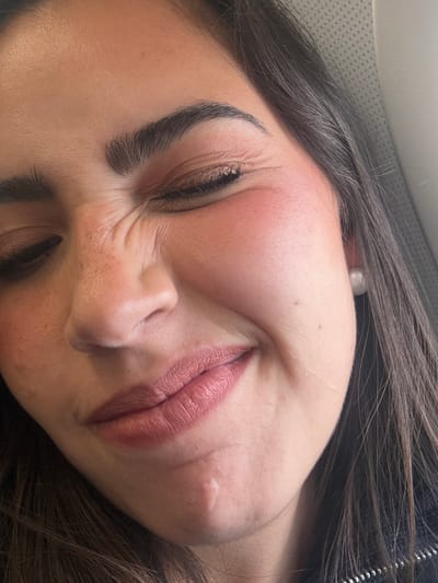 A close-up, angled view of a woman's face, likely taken indoors. The woman has dark hair and fair skin, with noticeable makeup, including defined eyebrows, blush on her cheeks, and lipstick. Her eyes are mostly closed, and her face is scrunched up, suggesting she is squinting or smiling widely. There is a small blemish or scar visible on her right cheek. The background is blurred, but appears to be a light-colored surface. There is no information about the location, or what city or country it was taken in.