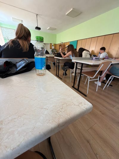 This image, taken in Slivnitsa, Bulgaria, depicts an indoor scene within what appears to be a classroom, study hall, or cafeteria. The photo is rotated 90 degrees clockwise.
In the foreground, the back of a person with blonde hair, tied into a ponytail, is visible. They are seated at a light-colored, marble-patterned table, wearing a dark top. On this table, a clear plastic water bottle with a green cap and a blue label is prominently placed, alongside a black wallet or small bag.
Beyond the foreground, several other students are seated at similar tables. A young man with dark hair, wearing a white t-shirt, is visible looking down at his table, with a purple bottle nearby. Another student with long, dark hair is also leaning forward over a table. The chairs have metal frames and wooden seats and backs.
The room features walls painted in two tones: a light green on the lower section and a lighter, possibly white or off-white, on the upper part. The floor is light brown wooden laminate. On the ceiling, rectangular fluorescent light fixtures are visible, one with a black device (possibly a speaker or sensor) attached. A blurred window and what appears to be a poster or board are in the background.
The students seem to be quietly engaged in individual activities, such as studying or using electronic devices. The text on the water bottle label is unreadable. The overall atmosphere suggests a typical daytime academic or informal learning environment.