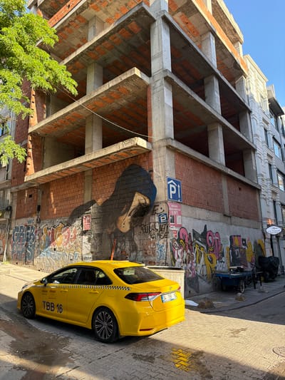 The image captures a street scene in Kadıköy, Turkey, featuring a yellow taxi with the license plate "34 TBB 16" parked on a cobblestone street. Behind the taxi, a building under construction with exposed concrete floors and brick walls is visible. The wall of the construction site is adorned with graffiti, including a large mural of a person's face. To the right of the construction site, a white building with an ornate facade stands. Further down the street, a blue cart and a dark tarp-covered object are present. The scene is illuminated by sunlight, casting shadows on the ground.