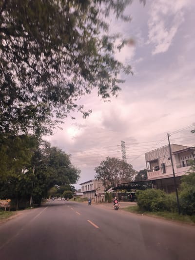 This outdoor image captures a road scene, likely taken from a moving vehicle, given the motion blur visible in the foreground trees and sky. The scene is set in Reuhattuha, Indonesia.
The primary subject is a paved road stretching into the distance, with faint yellow double lines marking the center and single yellow lines on the sides. Lush green trees line both sides of the road, particularly dense on the left, with their upper branches heavily blurred across the top and upper left of the frame. The sky is overcast with a mix of light and darker grey clouds, suggesting either an approaching storm, early morning, or late afternoon light.
On the right side of the road, several buildings are visible, some appearing residential and others possibly commercial or under construction with exposed frameworks. A tall metal lattice transmission tower and numerous power lines traverse the sky above these buildings. Further down the road, traffic includes several motorcycles; one with two riders (a person in a pink top and another in a dark top, both wearing helmets) is visible closer on the right, and other motorcycles and a white car can be seen in the distance.
On the left side of the road, partially obscured by trees, there's a sign with visible Indonesian text. The words "Selamat" and "PELAMINAN" are discernible, suggesting it might be a welcome sign related to a wedding or event, as "pelaminan" refers to a wedding dais. The general atmosphere is that of a typical, active road in a semi-urban or rural setting.