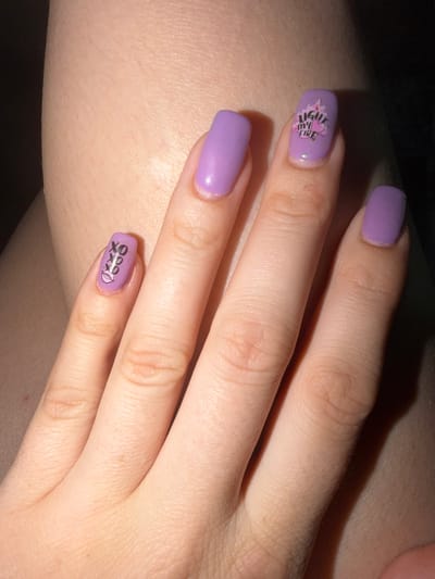 A close-up, high-angle shot showcases a person's left hand with purple-painted nails. The nails are decorated with small decals. The index finger has a decal of flames above a pink heart with the text "LIGHT MY FIRE" in black. The thumb has a decal of three "XOXO" phrases stacked vertically, with a pair of lips below them. The middle finger, ring finger, and pinky finger are painted solid purple. The hand rests against a person's skin, possibly an arm or leg, which appears to have fine hairs. The background is dark and out of focus. There is no identifiable information about the location in Fryazino, Russia.