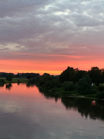 A river flows through a rural landscape under a dramatic sunset. The sky is a vibrant gradient of orange and pink, with streaks of gray clouds above. The trees lining the riverbank are dark silhouettes against the colorful sky, their reflections shimmering on the water's surface. In the distance, wind turbines are visible, suggesting the presence of infrastructure amidst the natural scenery. The water itself is calm, with gentle ripples disturbing its smooth surface. The scene conveys a sense of peaceful solitude and the beauty of nature at dusk. This scene is located in Rinteln, Germany.