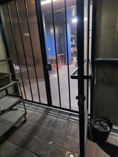 A perspective shot of a dark, metallic security gate partially open, revealing an interior space with blue walls. Several bicycles, some with red accents, are visible inside. To the left of the gate, a metal shelving unit holds a grey tub. The ground is paved with dark, textured tiles. A black pot filled with rocks and dirt sits to the right of the gate. The scene is outdoors, with shadows suggesting artificial lighting. The location is identified as being in Burbank, United States of America.